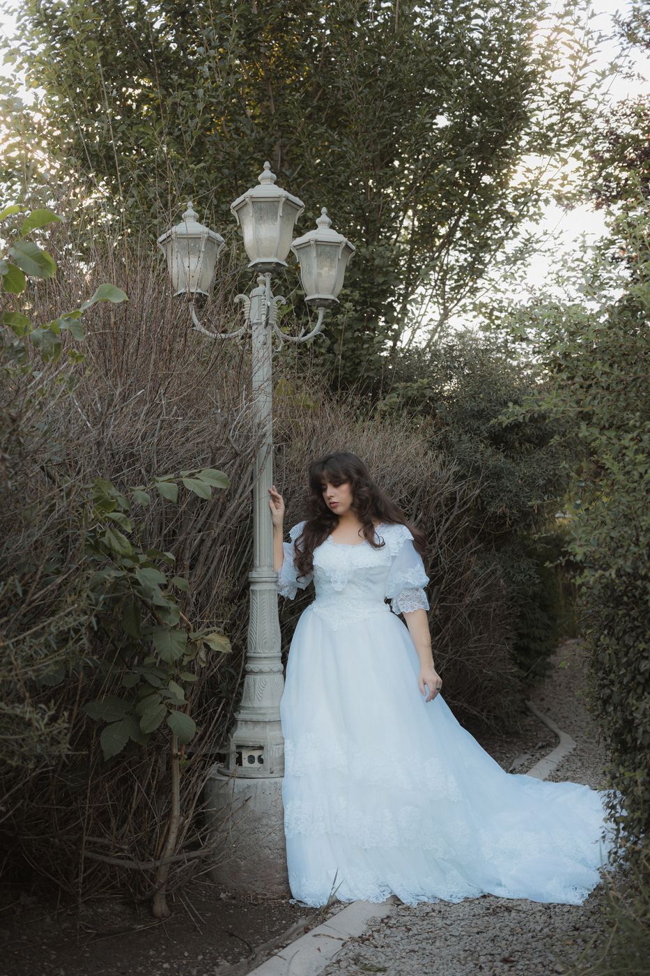 Bride in a pale blue off-the-shoulder gown standing beside a vintage white lamppost on a garden path lined with greenery