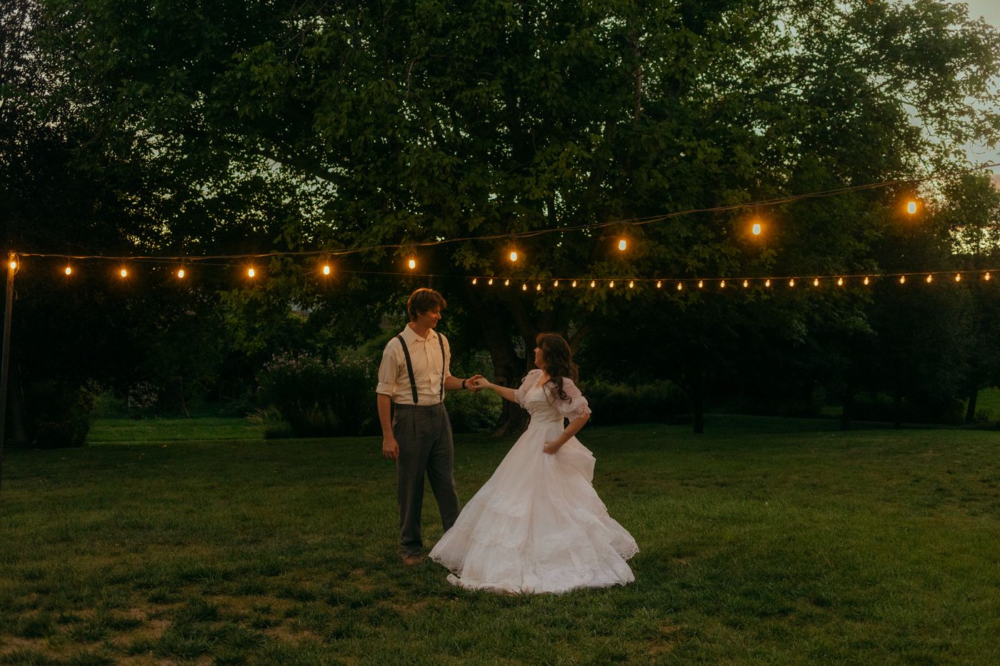 Couple dancing on the lawn in the evening under strands of bistro lights strung between the trees