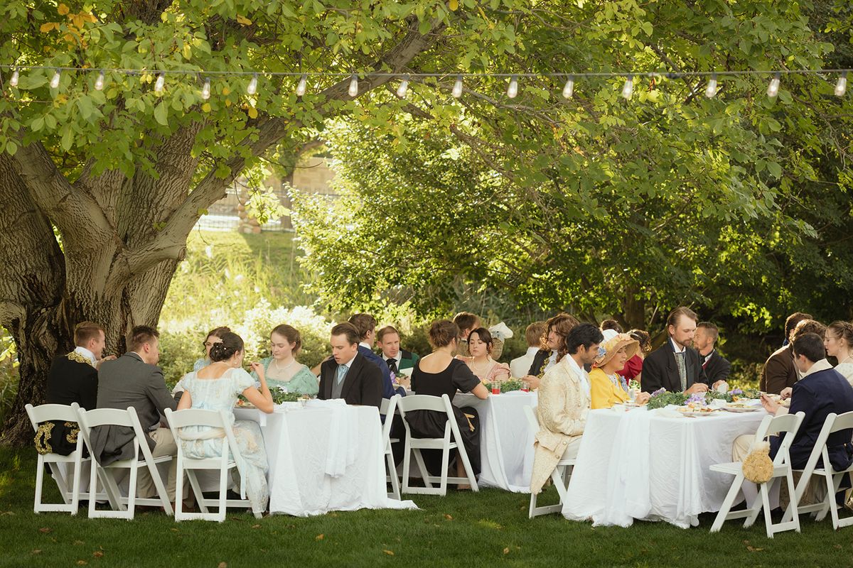 Guests seated at round tables with white linens under a large tree, with white canopy tents and string lights in the background