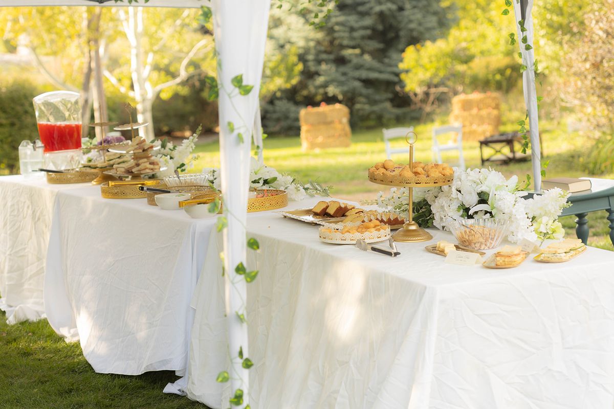 Buffet tables with white linens under a canopy, set with platters, a drink dispenser, and flowers, with hay bales on the lawn beyond