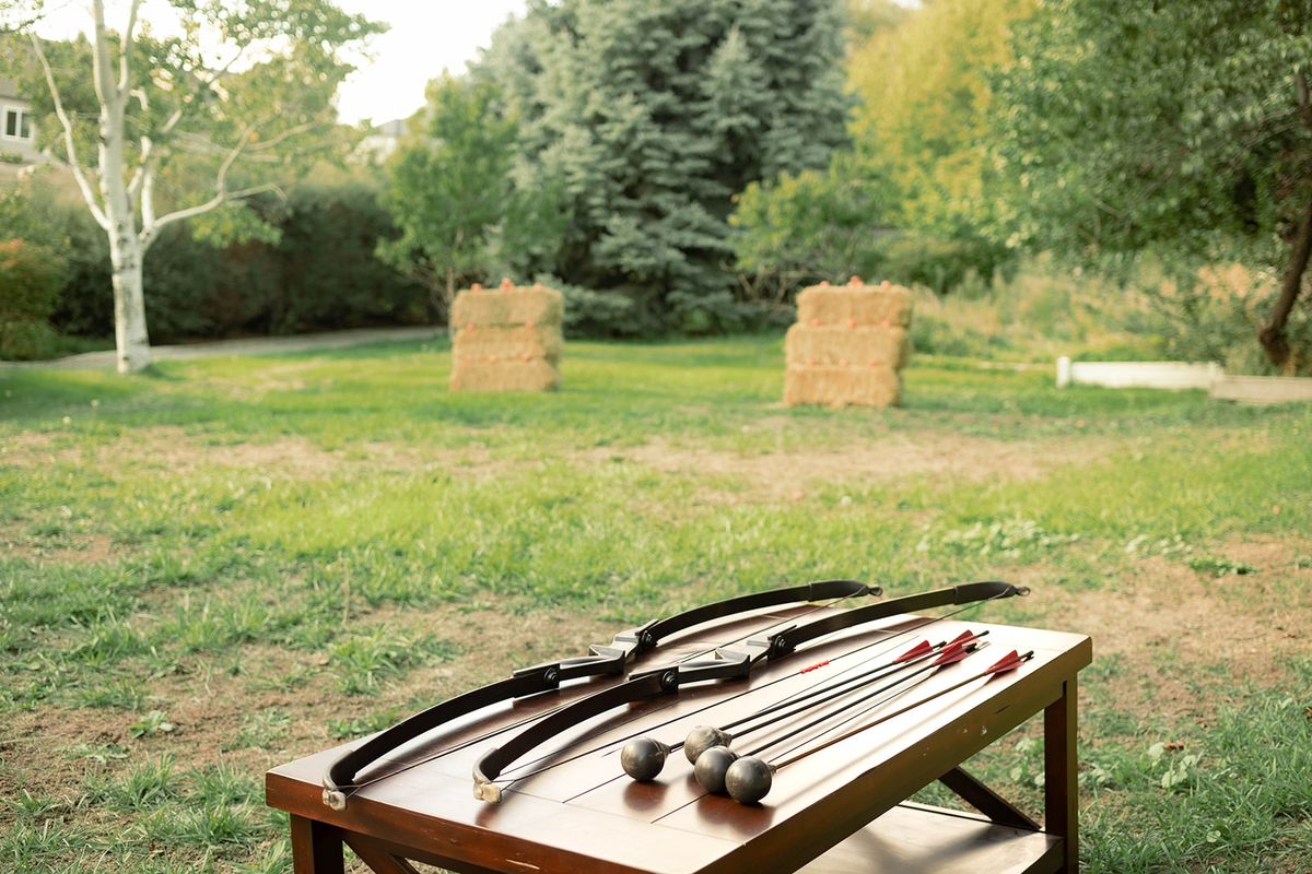 Wooden table holding archery bows and arrows on the lawn, with hay-bale targets and trees in the distance